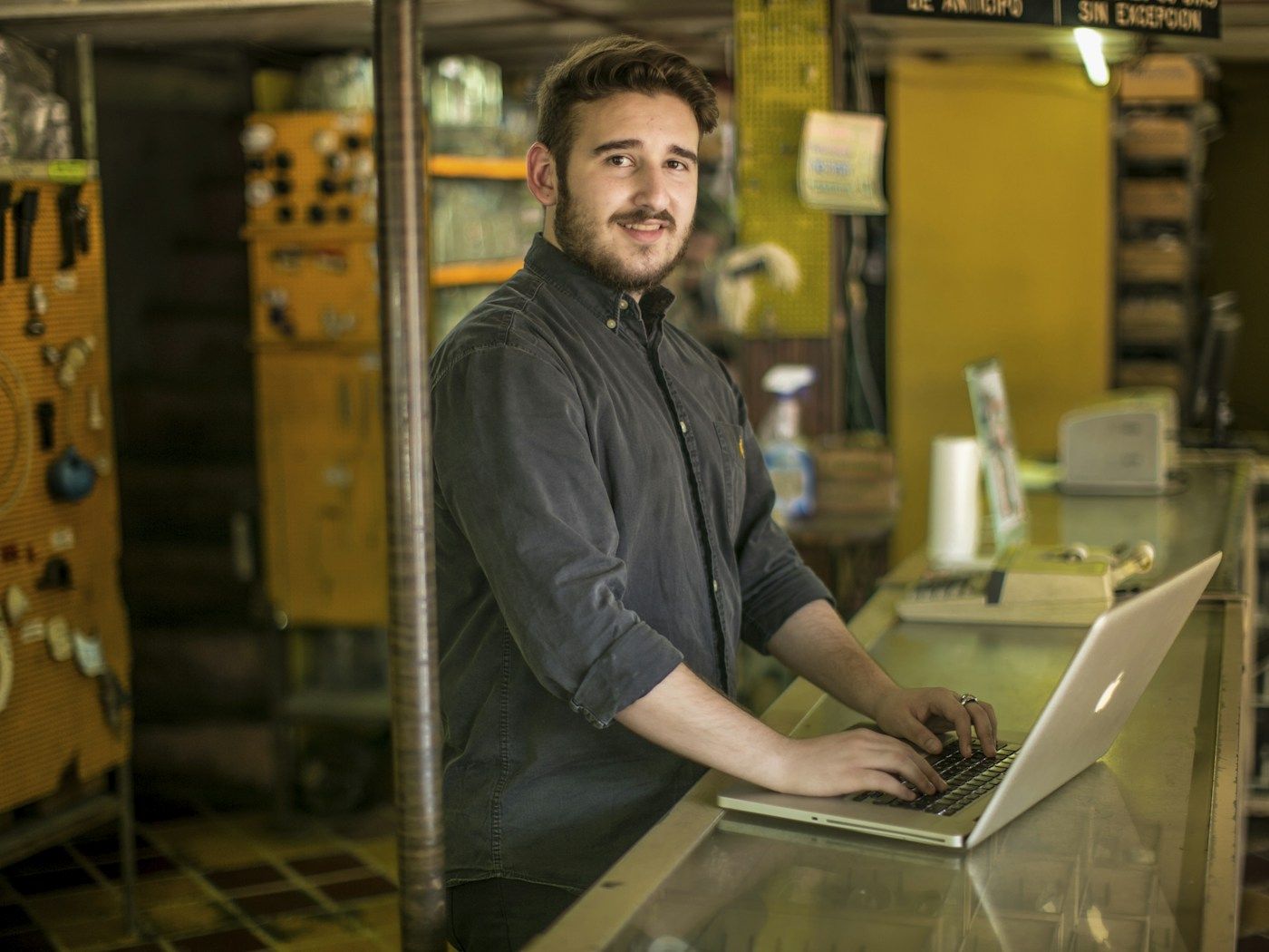 A shop owner smiles while working on a laptop at his glass counter, with tools hanging on the wall behind him