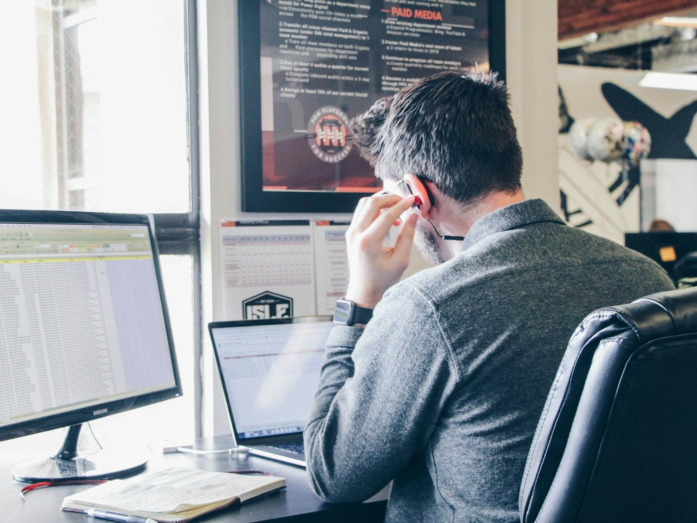 A man at a desk with a large spreadsheet on one screen and a laptop beside it