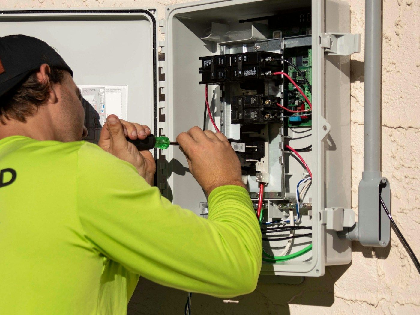 An installer in a bright work shirt wires an outdoor electrical panel with a screwdriver