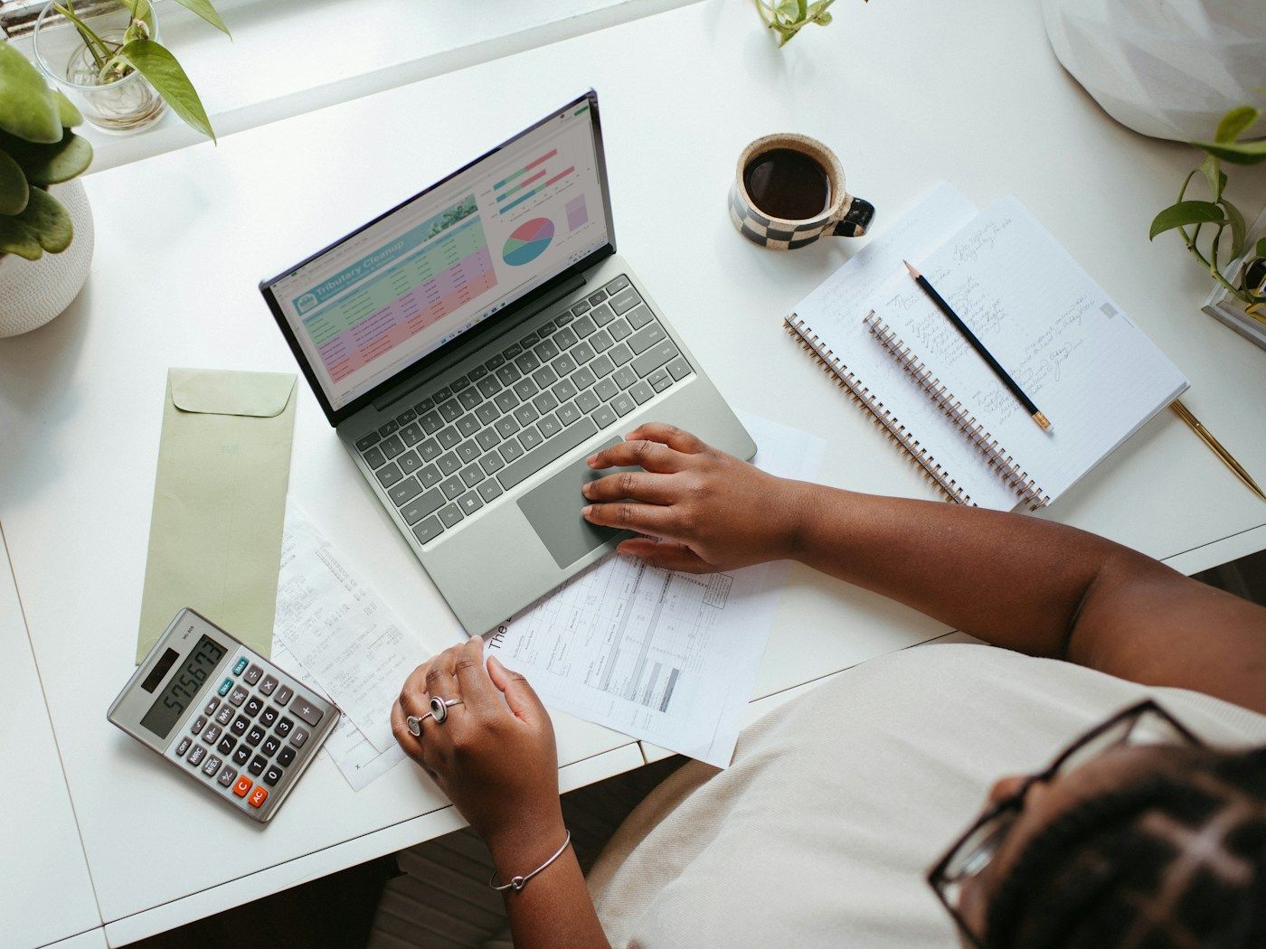 Seen from above, a person works on a laptop showing charts, with a calculator, receipts and a notebook on the desk