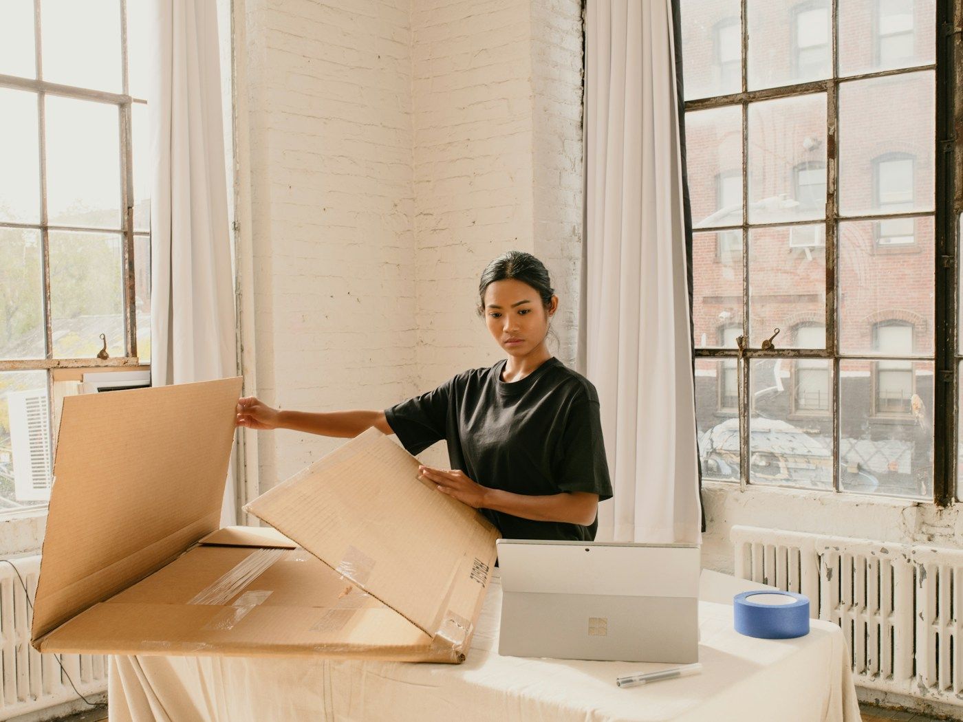 Photo: M. Cooper on Unsplash A woman folds a large cardboard shipping box at a table in a bright studio with tall windows