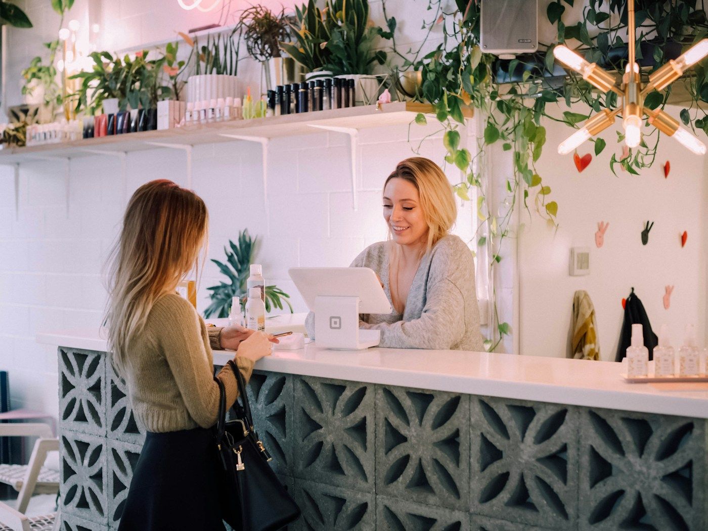 Photo: Christiann Koepke on Unsplash A customer stands at a plant-filled shop counter while the owner helps her at a tablet register